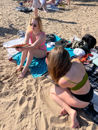 The image depicts a sunny day at a beach, with the sand clearly visible and marked with footprints and sand patterns. In the foreground, two young women are seated on beach blankets. The woman on the left, with blonde hair and sunglasses, is wearing a pink bikini and holding a decorative fan. She is looking to her right. The woman on the right has long, brown hair and is wearing a green bikini top and black bottoms. She is facing away from the camera and appears to be looking at her phone. Several items are scattered around them, including a bottle of drink, towels, bags, and clothing. In the background, other people are visible, also enjoying the beach. The scene suggests a casual outing at a public beach. This scene does not appear to be located in Ust'ye, Russia, as there are no identifying landmarks or cultural markers to suggest this location.