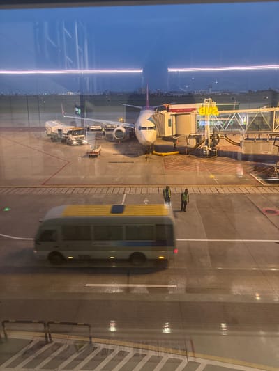 This image shows an airport tarmac at dusk, possibly in Xinwu District, China, as indicated by the accompanying metadata. A large passenger airplane is parked at a gate, connected by a jet bridge. Several airport vehicles, including a fuel truck and a baggage cart, are present on the tarmac. Two airport workers in high-visibility vests are standing near the aircraft. In the foreground, a bus is moving across the tarmac, its motion blurred, indicating it is in transit. The ground appears wet, suggesting recent rain or cleaning. The sky is a deep blue, transitioning into twilight, with lights from distant buildings visible on the horizon. The image is taken from an elevated perspective, likely from inside an airport terminal building, with reflections on the glass obscuring parts of the scene.