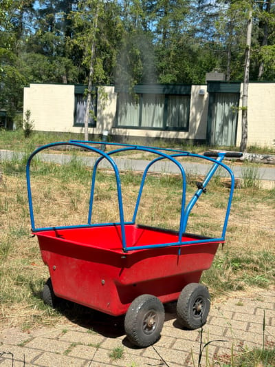 A bright red wagon with a blue frame stands on a paved surface in front of a low, cream-colored building with green trim and windows. The building is surrounded by dry grass and trees, suggesting a park-like or recreational setting in Heijen, Netherlands. The wagon has two large wheels in the back and appears to be unoccupied. The sky is clear and blue, indicating a sunny day.