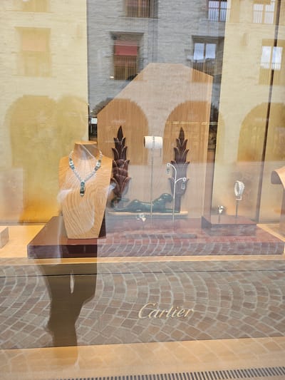The image shows a shop window display, likely a jewelry store given the items on display. Inside the window, there are several pieces of jewelry, including rings and necklaces, presented on wooden stands. One prominent display features a necklace with blue and clear stones on a light-colored wooden bust. The background of the display has a brick pattern and a visible "Cartier" logo, indicating the brand of the store, located in St. Moritz, Switzerland. Reflections of the exterior, including buildings with windows, are visible on the glass of the shop window. The lighting suggests it is daytime, possibly late afternoon, with sunlight casting shadows. The scene appears calm and static, typical of a retail environment.