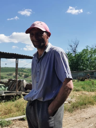 A man stands in a rural outdoor setting, possibly in or near Carbuna, Moldova, under a bright blue sky with scattered white clouds. He is wearing a light-colored, short-sleeved button-down shirt and dark gray sweatpants. A pink baseball cap is worn backward on his head. His left hand is in his pocket, and his right arm is bent at the elbow. The background shows a simple structure with a corrugated metal roof and wire fencing, suggesting an agricultural or storage area. Beyond this, rolling hills with green vegetation are visible, with some trees and what appears to be agricultural machinery in the distance. The ground in the foreground is dirt and dry grass.