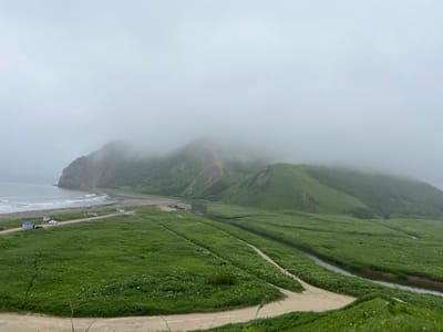The image depicts a scenic coastal landscape on a cloudy day, likely in the vicinity of Tikhoe, Russia. The foreground features a winding dirt road cutting through a vast expanse of lush green grassland dotted with small white flowers. Several cars are parked along another dirt road in the middle ground, near a small blue structure that appears to be a shelter or gazebo. This road leads towards a sandy beach and the choppy, gray ocean. In the background, rolling green hills rise up, partially obscured by thick fog that also covers the peaks of larger mountains in the distance. A small river or stream snakes through the valley between the hills, reflecting the muted light of the overcast sky. The overall atmosphere is serene and natural, with the fog lending a sense of mystery to the mountainous backdrop.