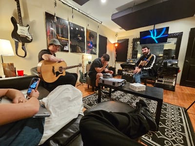 The scene depicts a music recording studio located in Hialeah, United States of America. Four individuals are present within the room. To the left, a male wearing a light-colored baseball cap and a dark shirt is seated in a gray upholstered chair, actively holding and appearing to play a light-wood acoustic guitar. Hanging on the wall behind him is a black and white electric guitar, and a floor lamp with an off-white shade illuminates the area. Three framed posters depicting musical artists are mounted on the wall. In the immediate foreground-left, partially obscured, an individual is seated on a dark couch, holding a smartphone over a silver laptop, observing the phone screen.

In the center-right of the image, a male in a dark t-shirt is seated on a low stool, resting his right hand against his forehead, looking downwards. Further to the right, a male with dark hair and a beard, wearing a black jacket with white stripes and blue jeans, is seated in a black office chair. He holds a small, brown stringed instrument, possibly a mandolin or ukulele, with a strap across his shoulder. Behind him, extensive audio recording equipment, including multiple racks of electronic gear and mixing consoles, is arranged. A flat-screen monitor displaying blue abstract patterns is mounted above this equipment.

A black coffee table occupies the center of the room, featuring a gray padded case and a small white box. The floor is covered by a black and white patterned rug over a wooden base. Dark acoustic treatment panels are affixed to the ceiling and walls, indicating a treated studio environment. The walls are light-colored, and the lighting within the room appears to be artificial and warm.