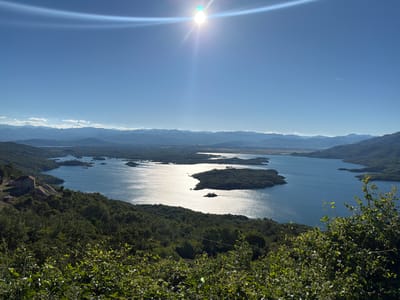 The image displays a scenic overlook of a large, expansive lake with numerous islands covered in lush green vegetation. The water is a deep blue, reflecting the bright sunlight, creating shimmering patterns across its surface. In the foreground, dense green foliage occupies the bottom portion of the frame, hinting at a natural, possibly mountainous environment. On the left side of the image, a stone building with a red roof is situated on a hillside overlooking the lake. The sky is a clear, brilliant blue, with the sun prominently positioned near the top center, causing a lens flare and lens artifacts. In the distance, a series of layered mountains fades into the horizon, some with touches of white clouds. A faint contrail from an aircraft is visible in the upper right quadrant of the sky. The overall impression is one of a vast, natural landscape with a sense of peacefulness and natural beauty, possibly located in or near Ponikvica, Montenegro, given the typical mountainous terrain and the presence of a large lake or reservoir. There are no people or active human interactions depicted in the scene.