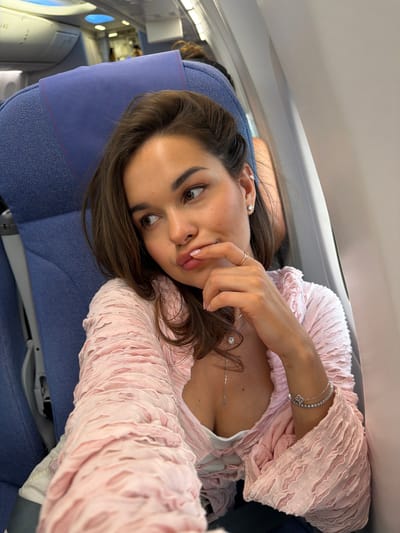 A woman is taking a selfie on an airplane. She is sitting in a blue airplane seat and leaning against the headrest. She is wearing a pink top, a silver bracelet, a ring, a necklace, and earrings. She is looking at the camera and touching her lips. The interior of the airplane is visible in the background, including the windows. The image was taken on an airplane but it is not possible to determine if the airplane is located in the Shunyi District, China.
