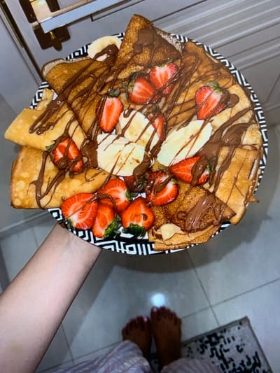 A plate of crepes topped with sliced strawberries, banana slices, and drizzled with chocolate sauce is held by a person's left hand. The plate has a black and white geometric pattern on its rim. In the background, the scene is out of focus, showing a kitchen counter with cabinets above. Below the plate, the tiled floor is visible, and a pair of bare feet wearing patterned fabric are visible in the lower right corner. The image does not provide any information to indicate the location in the city of Konstantinowka, Kyrgyzstan.