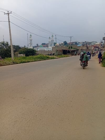 The image displays a daytime scene at Itakpe Junction, Nigeria, under an overcast sky. A wide, light brown road, appearing to be asphalt or compacted earth, extends from the foreground into the distance.

On the left side of the frame, a tall wooden utility pole with multiple overhead power lines stands. Below these lines, green vegetation, including bushes and small trees, borders the road, partially obscuring a concrete wall. In the mid-distance on the left, a prominent white mosque with two tall minarets and a central dome, all topped with green caps, is visible. To its left and slightly behind it, a grey telecommunications tower with multiple antennae is present. Beyond the mosque and vegetation, various buildings are clustered on an ascending hillside.

On the right side of the road, closer to the viewer, a dark motorcycle with a rider and a passenger, the latter wearing a green patterned garment, travels away from the camera. Further down the road, several other motorcycles and pedestrians are observed on the road and its unpaved shoulder. One pedestrian, dressed in a blue hijab, is visible near the right edge of the frame in the mid-distance. Structures with corrugated metal roofs, indicative of stalls or small shops, line the right side of the road further into the background, with additional buildings densely packed on the sloping terrain behind them.