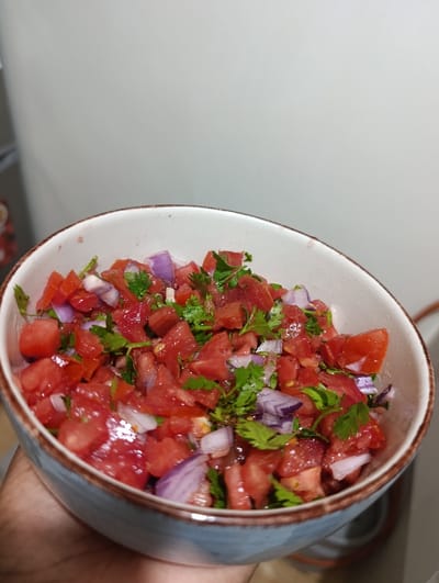 A close-up shot captures an individual's hand holding a light blue ceramic bowl with a rustic brown-rimmed edge. The bowl is filled with a vibrant, freshly prepared salad or salsa. The main ingredients visible are diced red tomatoes, chopped purple-red onions, and fresh green cilantro or coriander leaves, all mixed together. The ingredients appear moist and well-combined, suggesting they are ready for consumption.
The activity depicted is the presentation or holding of this food item, likely an appetizer or condiment. The background is a blurred, plain light grey or off-white surface, possibly a wall or part of an indoor setting, such as a kitchen or dining area. A faint, out-of-focus brown object, possibly a cable or part of an appliance, is visible in the lower right background. There are no specific time-of-day indicators, weather elements, or discernible emotions. No text is visible in the image. While the location context is Kahawa Wendani ward, Kenya, there are no specific visual cues within the image itself to confirm this geographical location.