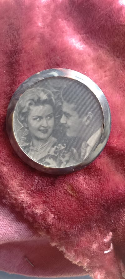 This media file shows a circular, silver-colored locket with a textured interior, resting on a textured, pinkish-red fabric. The locket contains a black and white photograph, or a depiction of a photograph, featuring two people. The faces of the individuals are visible, with one person's face prominently in the foreground, looking upwards and slightly to the left. The other person is positioned behind and above, with their face partially obscured, appearing to lean in. The background of the photograph within the locket has a striped pattern. The fabric beneath the locket is soft and appears to be a plush material. The overall scene suggests a personal item, possibly an antique, displayed on a textile surface. No specific time of day, weather, or clear location cues are discernible beyond the implied indoor setting. There is no visible text in the image.