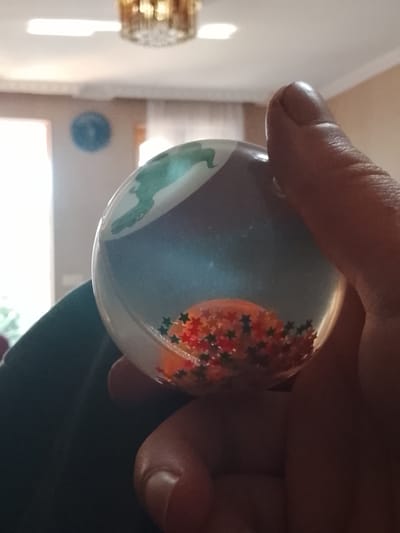 A close-up, slightly blurry, indoor photograph shows a person's thumb and index finger holding a clear, spherical object. The object appears to be a snow globe or similar decorative item. Inside the sphere, there is a green representation of what might be a dinosaur or a stylized dragon, and a scattering of colorful, star-shaped confetti at the bottom. The background suggests an interior setting with a ceiling fixture visible at the top, and a window with natural light entering from the left. A circular blue object, possibly a clock or decoration, is visible on the wall in the background. There is no visible indication of the location being in the city of Stalinisubani, Georgia.
