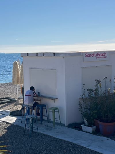 The image displays a beach scene in Civitavecchia, Italy, under a clear blue sky. A white, single-story kiosk-like structure with a flat roof is centrally positioned. Above an open serving window, a sign in pink lettering reads "Son of a Beach" with "give me a drink!" underneath. A wooden counter extends from the left side of the structure. A male individual, wearing a white polo shirt with red and blue stripes and dark shorts, is seated on a blue metal bar stool at this counter, facing inwards. His right hand is near his face, and he appears focused on an object on the counter. Three additional metal bar stools (one blue, two green) are arranged on a light-colored paved path in front of the counter. To the left of the structure, on a pebbled beach, a light-colored beach umbrella with a textured canopy is partially visible. The Mediterranean Sea, calm and blue, forms the background, with a large ship visible on the distant horizon to the left. Potted plants are positioned on the paved path to the right of the structure. The scene is brightly illuminated by natural sunlight.