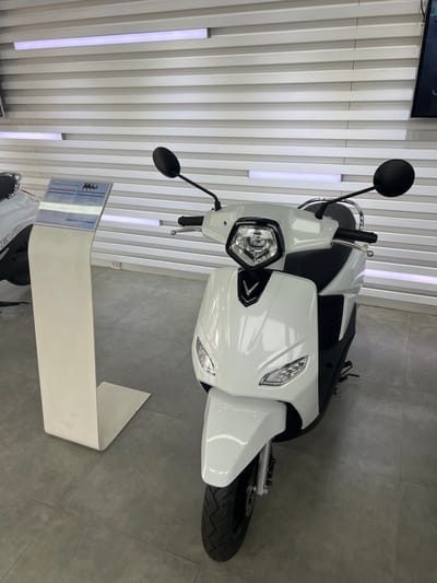 The scene is set indoors, likely within a modern VinFast dealership in Kon Tum, Vietnam. A white VinFast electric scooter, distinguished by a V-shaped emblem on its front cowl, a central LED headlight, and angular lower turn signals, is centrally positioned. It features black handlebars, circular black mirrors, and a black seat. The floor is covered with dark grey square tiles. To the left of the scooter, a white, angular display stand holds an information sheet detailing product specifications. Partially visible on the far left is the front portion of another white electric scooter, displaying "EVO" branding. The background consists of a white wall with horizontal ribbed panels, incorporating three integrated horizontal LED strip lights emitting a cool white glow. A double electrical outlet is visible on the lower left wall, and a dark screen is partially seen in the upper right corner. No people are present.