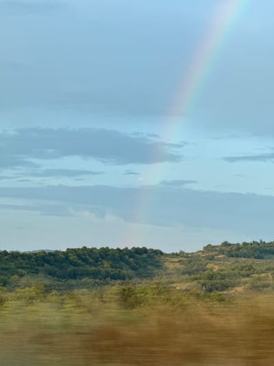 The image shows a landscape scene with a prominent rainbow stretching across a pale blue sky with scattered clouds. Below the sky is a rolling, tree-covered hillside. The foreground appears to be a blurred, brown field, suggesting the photo was taken while in motion. There are no people or man-made structures clearly visible in the image. The location is identified as Debelets, Bulgaria.