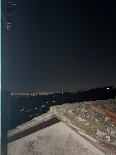 The image presents a night scene overlooking a body of water, potentially a lake or river. On the right, an aged stone structure, likely part of a building, is visible, featuring a decaying surface with exposed brick and mortar. In the background, on the opposite side of the water, a string of lights suggests a town or city stretching along the shoreline. The scene appears to be taken from a high vantage point, possibly from a building overlooking the water. The presence of the stone structure suggests an old or historic building in the vicinity. I cannot determine the specific location without additional information.