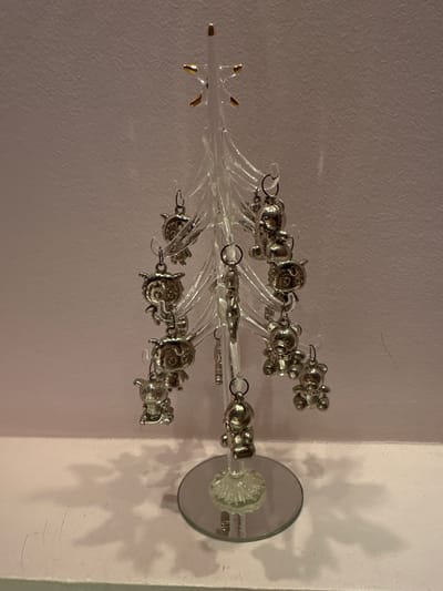 The image shows a small, clear glass Christmas tree with silver charms hanging from its branches. The charms appear to be of various animal shapes, including teddy bears and owl-like figures. The tree has a star on top and sits on a mirrored base. The background is a textured, pale pink wall, and a white surface is visible at the bottom of the frame, casting shadows from the tree and its ornaments. There are no people or discernible actions taking place. The setting is domestic or decorative, not specific to any particular location in Lobacheve, Ukraine.