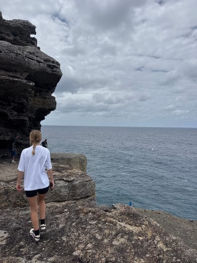 A woman with a blonde braid, wearing a white shirt and black shorts, stands near the edge of a rocky cliff overlooking a vast ocean. Her back faces the viewer. Behind her, rugged, dark rocks rise up. In the distance, other people are visible on the cliffside. Above, the sky is filled with textured, gray clouds, creating a sense of overcast weather. The ocean stretches to the horizon, meeting the cloudy sky.