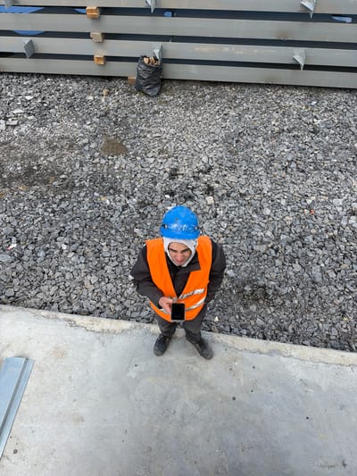 A construction worker stands on a concrete surface in Janowice, Poland. He is wearing a blue hard hat, an orange safety vest over a dark jacket, and dark pants. The worker is holding a smartphone in his hands, appearing to be looking at it. The environment is a construction site, with stacks of gray metal beams in the background, a pile of gravel covering the ground, and a black trash bag near the beams. A piece of metal construction material lies on the concrete in the foreground.