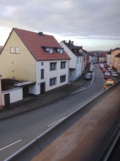 The image captures an elevated perspective of a residential street within Neunkirchen, Germany, under an overcast sky. The specific street name or precise location within the city is not discernible from the provided visual information.

In the immediate foreground, a weathered, brownish-grey balcony railing or ledge extends diagonally across the bottom right, partially obscuring the street below.

On the left side of the street, two prominent multi-story residential structures are visible. The building closest to the viewer on the far left exhibits a light yellow-beige shingle or panel facade on its visible side, topped by a steep red tiled gable roof. Two simple, rectangular windows are present on its upper level. Attached to its lower portion is a white, single-story extension with a dark brown door, possibly a garage or storage unit, featuring a flat, dark-colored roof extension.

Adjacent to this, directly fronting the street, is a three-story white building with a matching red tiled roof. This building features two dormer windows on the roof, a dark chimney stack, and multiple rectangular windows with dark frames, some equipped with external roller shutters. A dark-colored front door is centrally located on the ground floor.

The street itself is paved with dark asphalt, featuring a single continuous white line marking the center or edge. The sidewalk in front of the white building is paved with dark grey cobblestones or pavers. The street curves gently to the right and descends into the distance.

Numerous vehicles are parked along both sides of the street further down the curve. A dark grey passenger vehicle is parked on the left side, closer to the foreground. Further down the street, a yellow van is observed driving away from the viewer, its red taillights illuminated. Other parked vehicles include various sedans and vans, predominantly in lighter colors, lining the street compactly.

The buildings lining the street as it recedes into the background are multi-story residential units, exhibiting a variety of pastel facades (e.g., light blue, cream) and traditional European architecture. The distant horizon reveals more urban development under the uniformly grey, cloudy sky. No individuals are visible within the scene, and no explicit actions or interactions are occurring. The environment suggests a typical, quiet European town setting.