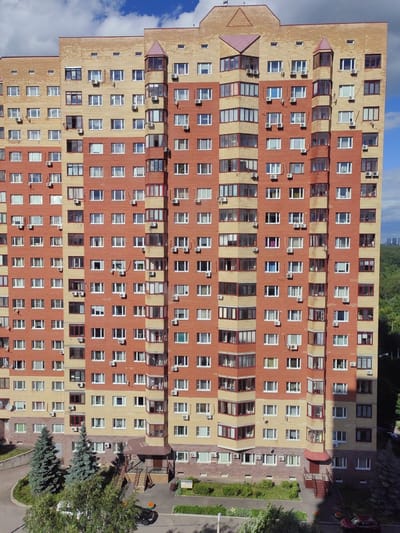 An aerial drone shot captures a large, multi-story apartment building made of brick in Putilkovo, Russia. The building features a combination of red and yellow bricks, with numerous windows and balconies. Two conifer trees are visible in front of the building, along with a paved area with an entrance to the building and stairs. A black car is parked on the paved area, and a red car is parked on the right side of the frame. The sky is partly cloudy.