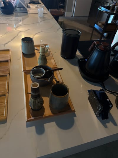 This image depicts a countertop display of tea-making accessories, likely in a cafe or home setting, located in North Miami Beach, United States of America. The scene is dominated by a marble-patterned countertop, reflecting ambient light.
On the left side of the frame, a stack of wooden trays is visible. In the center, a wooden tray holds a collection of matcha tea preparation items. These include a blue ceramic cup, a bamboo whisk (chasen), and a small sieve with a wooden handle resting over a dark ceramic bowl. Further down the tray are another bamboo whisk and a dark green ceramic cup.
To the right of the tray, a black electric kettle with a wooden handle sits on a black charging base. Adjacent to the kettle is a black cylindrical container, possibly for tea leaves or storage. In the foreground on the right, a black marble-patterned napkin holder with dark blue napkins is positioned on the countertop. The background is blurred, showing glimpses of shelves with various items and a stool. The overall impression is a neatly arranged setup for preparing and serving matcha tea.
