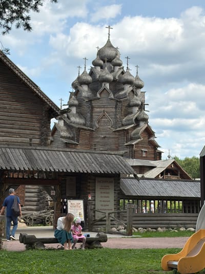The image depicts a scene at a wooden architectural complex, likely a museum or historical site, in Nevskii Parkleskhoz, Russia. A prominent, intricately designed wooden church with multiple onion domes and crosses dominates the background. In the foreground, a man in a blue shirt and jeans walks away from the camera towards the left side of the frame. To the right, a woman in a white top and a teal skirt, and a young girl in a pink dress, are seated on a log bench. The girl is looking down at her hands, while the woman's head is also bowed. Behind them, a wooden fence encloses an area with more wooden structures. Signs with Russian text are visible, one of which appears to be a menu for excursions. The environment is lush green grass, with trees and a cloudy sky overhead. A yellow slide is visible on the far right of the frame, suggesting a recreational area.
