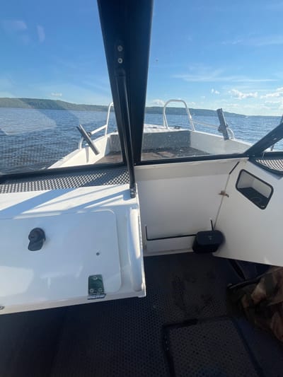 The image shows the interior of a boat navigating on a body of water, with land visible in the distance. The boat is white with black trim and has a windshield. In the foreground, there is a white storage compartment with a black latch and a metal hinge. The floor of the boat is covered with a black, textured material. A black object with the word "PUSH" is visible on the right side of the boat. The water is calm with gentle ripples, and the sky is blue with scattered white clouds. The land in the distance appears to be covered in green trees. Based on the context, the boat is likely located on the river near Siukterka, Russia, a village situated on the banks of the Tom River in the Kemerovo Oblast.