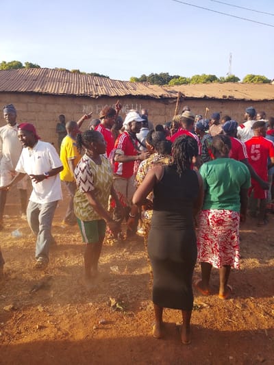A group of people gathers outdoors. The majority are dressed casually in various colored shirts and dresses. A wall made of mud bricks and topped with corrugated metal sheets is visible in the background, along with a tower. The ground is dirt with scattered debris. Some people are wearing hats or head coverings. The overall atmosphere suggests a community gathering or celebration.