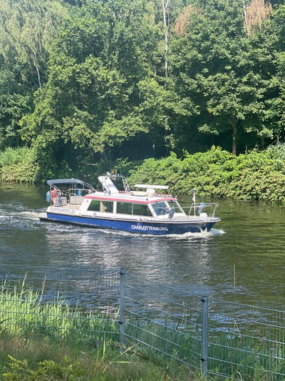 A police boat, marked "SCHIFFAHRTSPOLIZEI CHARLOTTENBURG," travels down a waterway, creating a wake. The boat is white with blue and red trim, and has a covered seating area at the stern. It is equipped with navigational equipment. The waterway is bordered by lush green trees and vegetation on both sides. In the foreground, a metal fence with a wire mesh runs along the grassy bank. The sky is clear and the sun is shining. The scene appears to be in a natural setting within or on the outskirts of a city.