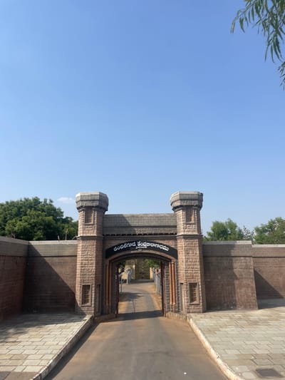 A low-angle, horizontally-oriented photograph depicts the entrance gate to the District Central Jail in Anajpur, India. The gate is constructed of tan stone or brick with darker gray accents, featuring two towers flanking a central archway. A sign, likely in Telugu script, spans the arch. Beyond the gate, a road or walkway extends into the prison complex. The sky above is clear and blue, with foliage visible at the top left and behind the gate structure. A paved area is on the right side of the road.
