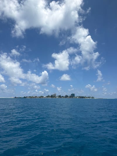 The image captures a wide-angle view of the island of Gulhi, Maldives, observed from a body of open ocean water under a clear blue sky. The lower half of the frame is dominated by deep blue ocean water, characterized by a textured surface of small ripples and waves.
The island, identified as Gulhi, stretches horizontally across the mid-ground, presenting a relatively flat profile. Its shoreline, visible as a light-colored strip, gives way to a densely vegetated interior with green trees and foliage. Numerous man-made structures are present on the island. Towards the left, smaller buildings and what appear to be docked boats or vessels are visible near the water's edge. In the central and right portions of the island, multiple multi-story buildings are apparent, including a prominent light-colored structure and a darker, rectangular building. Various other smaller buildings and utility poles or masts are interspersed throughout the visible landmass.
The upper half of the image features a bright blue sky filled with numerous distinct, white cumulus clouds of varying shapes and sizes, indicating fair weather conditions. No specific individuals or active interactions are discernible within the frame.