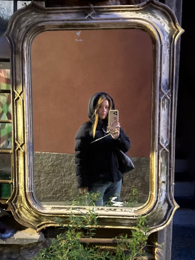 The image displays a female subject captured in a self-portrait reflection within a large, rectangular, ornate mirror, positioned outdoors in Monterosso al Mare, Italy. The female is centered in the mirror, wearing a black puffer jacket with the hood raised, blue jeans, and carrying a black shoulder bag on her left side. Her blonde hair is visible beneath the hood. She holds a light-colored smartphone, featuring a pink flamingo graphic on its case, in her right hand, angled towards the mirror. The mirror's frame is heavily weathered, exhibiting chipped details on its gilded or brass-colored, curvilinear, and geometric design. Green foliage is growing at the base of the mirror, partially obscuring its lower edge. The background reflected in the mirror consists of a reddish-brown wall above a textured grey stone or concrete section. To the reflected left, a metal grate with a multi-colored, patterned background is visible. The reflected area to the right is dark, suggesting a shadowed space. The scene is illuminated by an artificial light source.