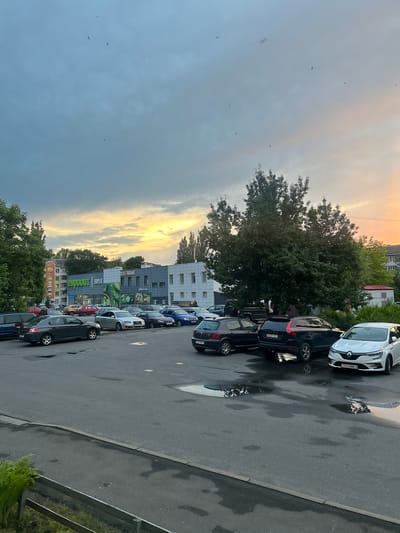 The image shows a parking lot in front of a supermarket named "Euroopt Super" in Mazyr, Belarus. The sky is overcast with the sun setting, casting a warm glow on the clouds and illuminating some parts of the sky with orange and yellow hues. There are several cars parked in the lot, including a white Renault, a black Volvo, and a dark grey sedan in the foreground. Some puddles of water are visible on the asphalt, reflecting the surrounding environment. In the background, there are several buildings, including the supermarket and residential buildings. Trees are also scattered throughout the scene. A few birds are visible in the sky. The scene appears to be calm and ordinary, typical of an urban environment during the evening.