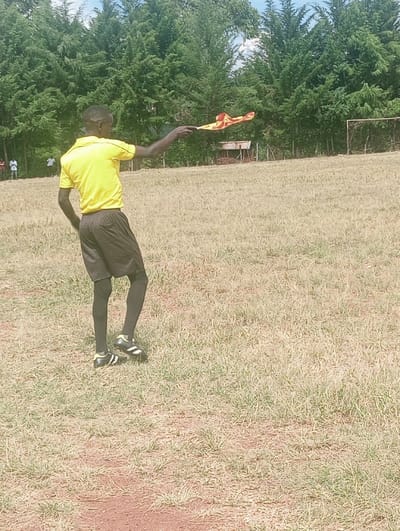 An individual, identified as an assistant referee, stands on a dry, sparsely vegetated sports field in Lwandeti ward, Kenya. The person, viewed from the back and slightly to the left, wears a yellow short-sleeved shirt, black shorts, black athletic socks or leggings, and black-and-white studded athletic shoes. The right arm is extended horizontally, holding a red and yellow checkered flag on a stick. In the background, a line of dense green trees borders the field. To the far right, a metal goalpost with a net is visible. A rustic wooden structure is partially discernible among the trees. The ground consists of dry, brownish-green grass with visible patches of reddish-brown earth. The scene is illuminated by natural daylight.