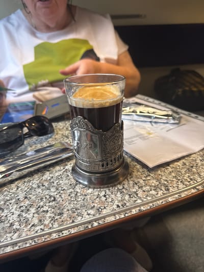 In Novye Biriuli, Russia, a close-up shot shows a glass of dark liquid topped with a frothy cream, held in a decorative silver holder with intricate engravings. The glass is placed on a speckled granite-like surface. To the left, a pair of black sunglasses rests beside a metallic object and what appears to be a business card. In the background, out of focus, a person wearing a white t-shirt with a green graphic is visible, along with some papers and a dark bag. The lighting suggests an indoor setting, possibly a cafe or a train compartment, with the focus on the drink and its accompaniments.