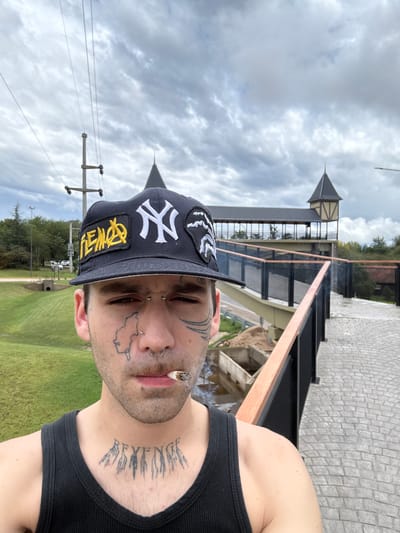 A selfie depicts a man outdoors, likely on a bridge or elevated walkway, smoking a cigarette. He is wearing a black tank top and a black New York Yankees baseball cap. He has multiple facial tattoos and piercings. The background shows a partly cloudy sky, trees, grass, and a building with a distinctive turret. The location is identifiable as Villa General Belgrano, Argentina, based on the architectural style of the building in the background, which resembles traditional German architecture common in that town. The man is holding the phone or camera and taking the selfie. The scene appears to be during daylight hours.