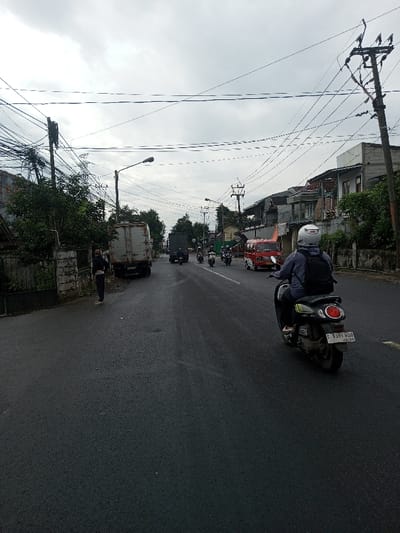 The depicted scene is an urban street, identified as being in the city of Kamji Na-Yekha, Bhutan. However, a visible license plate 'F 6381 HSO' on a scooter and the architectural style are characteristic of urban environments in West Java, Indonesia.
The scene is viewed on an overcast day. The asphalt road is dark and appears damp. In the foreground, a person wearing a white helmet, a light-colored jacket, and a dark backpack is riding a dark scooter away from the viewer. The scooter has a red taillight and its rear license plate displays 'F 6381 HSO' with '04-28' below.
On the left side of the road, a second individual in dark attire and a face mask stands near a concrete wall, facing the street. Behind this person, the rear of a light-colored, soiled box truck is stationary or parked. A utility pole immediately behind the truck is heavily strung with numerous overhead electrical and communication cables. Further left, residential structures and green foliage are visible.
In the mid-ground, a dark truck, followed by a procession of at least four scooters/motorcycles, is traveling away from the camera. A red minivan or microbus is also moving in the same direction on the right side of the road. The right side of the street is bordered by a low wall and a series of multi-story buildings, consistent with residential or commercial properties, interspersed with additional utility poles and overhead wiring.