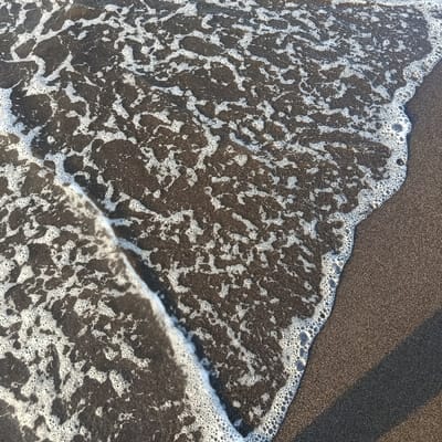 The image shows a close-up view of a sandy beach in Arjasa, Indonesia, at coordinates -7.71 latitude and 114.118 longitude. The receding tide has left a pattern of white foam and bubbles on the dark gray sand. The foam forms a distinct line against the sand, creating a clear boundary between the wet and dry areas of the beach. The sand appears to be fine-grained and compact, with subtle variations in texture. The image captures a moment of calm and tranquility on the beach, showcasing the natural beauty of the coastline.