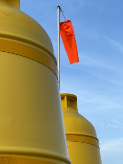 The image shows two large yellow cylindrical objects, possibly buoys or industrial tanks, positioned in close proximity. They are both adorned with a reflective yellow band around their circumference. In the background, a silver pole stands erect, from which an orange windsock is suspended, indicating wind direction. The sky is a clear blue with a few wispy white clouds. The setting appears to be outdoors, possibly near a body of water or in an industrial area. No people are visible in the frame. The overall impression is one of industrial or maritime equipment against a natural backdrop. The location of Terhillion, Ireland is not discernible from the image itself.