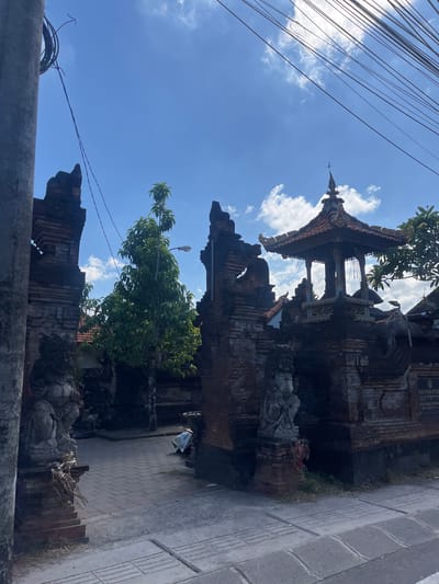 A low-angle, medium shot captures a traditional Balinese temple entrance with weathered brick structures and ornate carvings under a bright blue sky dotted with scattered clouds. The gate features two imposing stone statues of guardians, one on each side, at the entrance. A lush green tree stands tall between the gateway pillars. Overhead, a network of dark electrical wires crisscrosses the sky, suggesting an urban or semi-urban setting within Denpasar Barat, Indonesia. The paved walkway leads into the temple courtyard, where a tiered, pavilion-like structure with a red tile roof is visible in the background. A street sign is also partially visible on the right side of the image.