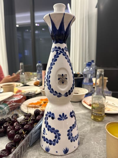 This is a medium-angle, eye-level, close-up shot of a decorative bottle with a blue and white floral pattern, positioned on a grey marble-like surface. The bottle is ornate, with a flared top and a detailed blue floral design covering its body. A small, silver, four-petaled flower adorns the front of the bottle. In the foreground, to the left of the bottle, is a clear plastic container filled with dark red cherries.
Behind the bottle, the background is blurred, suggesting a gathering or meal. Several plates of food are visible, including what appears to be toast with orange roe, and another plate with red sauce, possibly ketchup. There are also several bottles of water and a glass bottle with a yellow liquid and a label in Chinese characters, likely containing an alcoholic beverage. A disposable paper cup filled with a yellow liquid is visible on the right side. In the far background, people are dimly visible, with hands holding mobile phones, and windows with reflections of interior lights. The overall impression is that of a casual social gathering, potentially a meal or party, taking place indoors, possibly in a dining or living area. The location is specified as Qingcheng District, China.