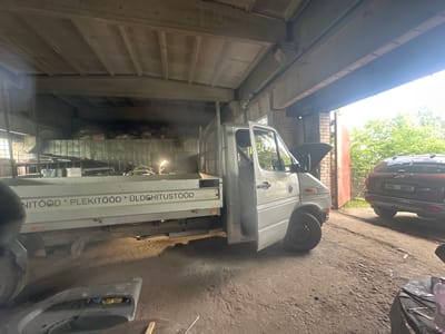 This media file depicts an indoor scene, possibly a garage or workshop, in Kohtla-Järve linn, Estonia. The setting appears somewhat cluttered and utilitarian, with a concrete floor and a corrugated metal roof supported by exposed beams. Natural light streams in from the right side, suggesting it's daytime.
The primary subjects are two vehicles. On the left is a white truck, a Mercedes-Benz Sprinter, with its driver's side door open and the hood up, indicating maintenance or repair work. The side of the truck's cargo bed has text printed on it: "NITÖÖD * PLEKITÖÖD * ÜLDEHITUSTÖÖD". To the right of the truck is a reddish-brown SUV, likely a Ford, parked facing away from the camera. The SUV has a license plate that reads "914 SRX".
Other objects visible include what appears to be a car bumper or fender on the ground in the foreground, and various tools and materials are scattered around the workshop. In the background, stacks of items covered with tarps or plastic are visible, as well as some brickwork on the right wall. The overall atmosphere suggests an active workspace. The lighting and shadows indicate it is likely daytime, and the presence of trees visible outside the open garage door suggests a temperate outdoor environment. No people are clearly visible in the frame.