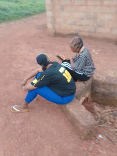 This outdoor photograph depicts two individuals, likely women, seated on rough stone or concrete blocks on a reddish-brown earth ground in Ugwolawo, Nigeria.

The individual in the foreground is viewed from the back and side, wearing a black t-shirt with yellow detailing, notably the number "18" prominently displayed. She pairs this with bright blue leggings and light brown flip-flops featuring white straps. A patterned head covering is also visible. Her posture suggests she is resting or looking down, with her hands clasped.

Behind her, a second individual is seen from a similar angle. She wears a horizontally striped shirt, possibly black and white or grey, and dark athletic pants with white stripes along the side. She also has a patterned head covering. This person appears to be engaged with a small electronic device, likely a smartphone.

The background shows a simple wall constructed from light-colored concrete blocks and blurred green foliage, indicative of a natural, perhaps rural or semi-rural, environment. The ground is unpaved soil. The lighting is soft and even, suggesting daytime conditions, possibly late afternoon. No strong emotions are discernible from their postures. The scene conveys a sense of quiet relaxation or casual activity.