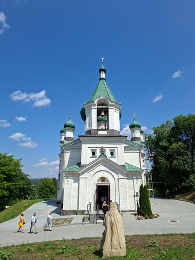The image depicts the exterior of a white church with green domes, located in the city of Condrița, Moldova. The church features a central bell tower with a large bell and a cross at its apex. Several smaller domes with crosses are also visible. The church entrance is open, and a few people are seen entering and exiting.
In the foreground, a rough-hewn wooden sculpture stands. To the left of the church, a paved pathway leads up to the entrance, with a grassy slope beside it. Three individuals are walking on the path, dressed in casual attire. To the right of the church, a tall evergreen tree stands next to a small flower bed with a cross. Beyond the church, lush green trees form the background, with a hilly landscape visible in the distance. The sky is a clear blue with a few scattered white clouds. Scaffolding is erected near the side of the church.