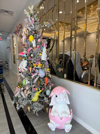 The image depicts the interior of a commercial establishment in Naples, United States, likely a nail salon or spa. The central focus is a large, frosted artificial tree, extensively decorated for Easter with numerous white and pastel bunny figures, multi-colored eggs (yellow, pink, blue, green), carrot ornaments, floral accents, and various ribbons. Positioned directly to the right of the tree is a white rabbit statue with blue eyes, emerging from a pink cracked egg adorned with white polka dots.
A modern, reflective gold-paneled partition occupies the mid-ground on the right, separating the primary display area from a section featuring multiple white and black pedicure chairs. Through the gaps in these panels, one female individual is visible seated in a pedicure chair, wearing sunglasses on her head and a light-colored top, with her feet immersed in a basin.
The environment features a white ceiling with square air vents and recessed lighting, contrasting with a high-gloss floor composed of alternating grey and black diagonal stripe tiles. Reflections of elaborate, multi-tiered chandeliers are visible in the gold panels. An "EXIT" sign with a red arrow is affixed to a wall in the far left background, near a hallway entrance.