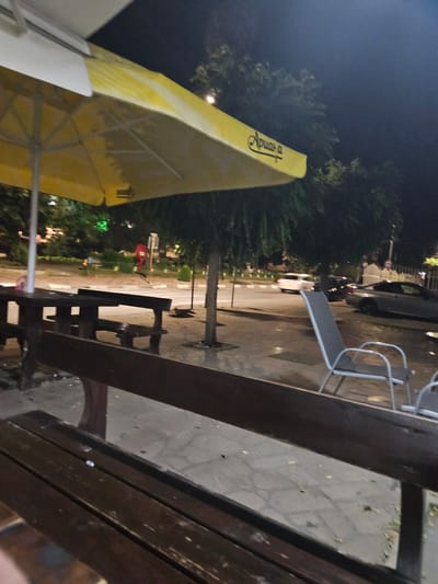 The image depicts an outdoor seating area at night, likely a cafe or restaurant, in the city of Byaga, Bulgaria. A large yellow umbrella provides shade over wooden picnic tables and benches. In the background, trees line a street with parked cars and a white vehicle is visible driving away. A silver sedan is parked on the street. A grey metal chair with a light-colored seat and back is positioned near the tables. The ground is paved with cobblestones, and fallen leaves are scattered across the surface. The overall impression is one of a quiet, possibly post-dinner scene, with the artificial light from the street and surrounding buildings illuminating the area.