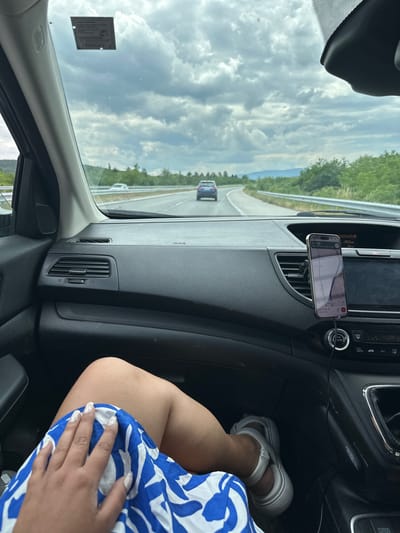 The image provides a passenger's perspective from inside a car traveling on a highway during daytime. The sky is largely overcast with scattered clouds.
In the foreground to the right, a person's lower body is visible. They are wearing a blue and white patterned outfit and white PUMA-branded sandals on their feet. Their left leg is casually propped up, and their hand, adorned with white, patterned manicured nails, rests on their lap.
The car's dark-colored dashboard occupies the middle ground. A smartphone is mounted horizontally on one of the central air vents, displaying a navigation application. The screen shows the time "22:20", a remaining distance of "222.0km", and a speed of "10.0km/h". Some Cyrillic text is visible on the map interface. A charging cable is connected to the phone. Below the phone, various car controls and buttons are visible, including "A/C", "FRONT", "AUTO", and a "POWER OUTLET 12V 180W".
Beyond the car's interior, a multi-lane highway stretches into the distance, flanked by green trees and what appear to be mountains in the background. A white guardrail runs alongside the road. Another car, dark blue in color, is visible ahead on the highway. A small, rectangular sticker, likely a vehicle registration or vignette, is affixed to the upper left side of the windshield, though its text is illegible. The scene depicts a relaxed journey through a natural landscape near Djerman, Bulgaria.