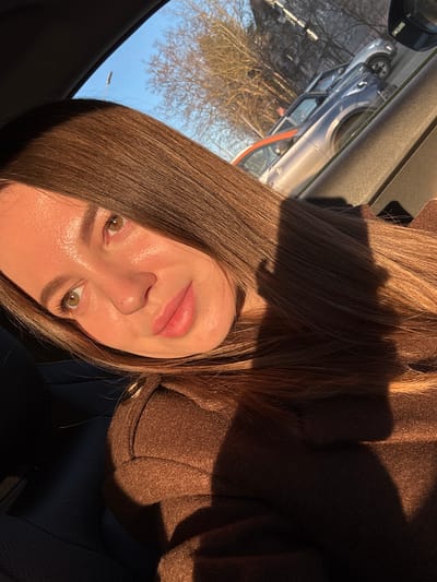 A medium close-up photograph shows a woman with long brown hair and green eyes in the interior of a vehicle. Her face is illuminated by sunlight. She is wearing a brown coat. Outside the vehicle, there are trees and a silver car parked on the street. Based on the provided location, this is Likino-Dulyovo, Russia. The photograph appears to be taken inside a car looking out towards the street, with the woman positioned inside.