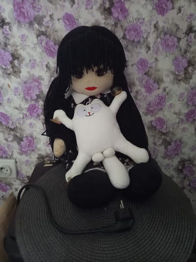 A doll with black yarn hair, large eyes, and red lips sits on a gray, circular surface with a coiled black cord and plug. The doll holds a white plush cat with a smiling face. The background is a floral wallpaper with purple and gray flowers. A white electrical outlet is visible on the left side of the image. The scene is indoors.