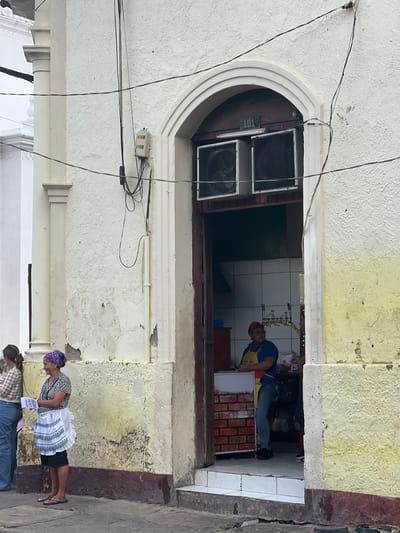 In León, Nicaragua, a street scene unfolds with two women and a man visible outside and inside a building. On the left, a woman with dark hair pulled back stands wearing a patterned top and blue jeans, holding some papers. Beside her, a woman wearing a purple headscarf, a black and white patterned shirt, and a white apron over dark shorts, smiles as she holds papers. Their attire suggests a casual or working environment.

The building features a textured, off-white facade with peeling paint near the bottom, accented by a dark, reddish-brown base. An arched doorway, painted dark brown, leads into the interior. Above the doorway, the number "101" is visible in a small sign, and two large, industrial-looking fans are mounted. Inside, behind a counter with a brick-patterned facade, a person wearing a blue shirt, red cap, and a yellow apron is visible. They appear to be working or attending to customers. The interior shows tiled walls and various items, including what looks like food packaging. The overall atmosphere suggests a commercial establishment open to the street. Wires are strung across the building's exterior, contributing to the urban streetscape.
