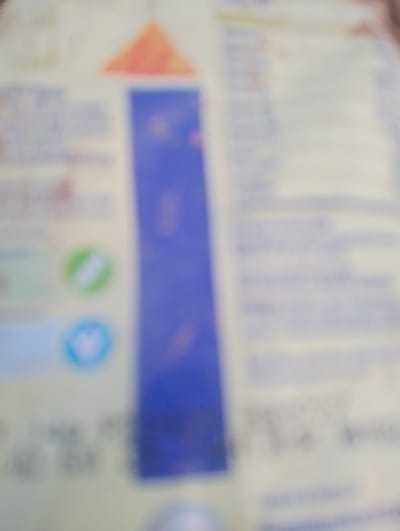 The image is a blurry, close-up, angled photograph of what appears to be packaging for a food product. The dominant colors are white, light yellow, and a large vertical blue stripe on the right side. There are some faint graphic elements, including what looks like a red triangle at the top left, and two circular icons below it. One circle is green with a white diagonal stripe, and the other is light blue with a white symbol inside that resembles a heart or a stylized "V". Text is visible in a blue font, but it is largely unreadable due to the blurriness. The background is a plain, light-colored surface. The image does not contain any discernible information about a location in Rahwol Chom, Nigeria, or any people or specific actions.